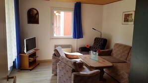 A living room with a TV, a sofa, a chair, a table, and a window with curtains. - Quietly located bungalow with a view of the dam (Zeulenroda-Triebes)
