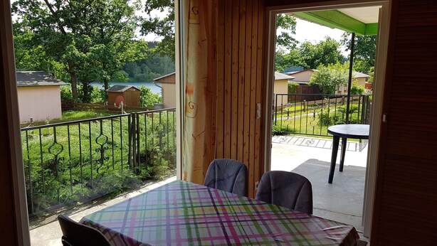 A balcony with a table and chairs, a view of a garden, and a wooden door. - Quietly located bungalow with a view of the dam (Zeulenroda-Triebes)