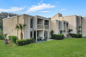 Un edificio de dos pisos con techo plano, un balcón en el segundo nivel y un pequeño patio amoblado.