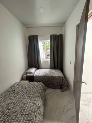Ein kleiner, gemütlicher Raum mit zwei Betten, einem Fenster mit Vorhängen und einer Tür.