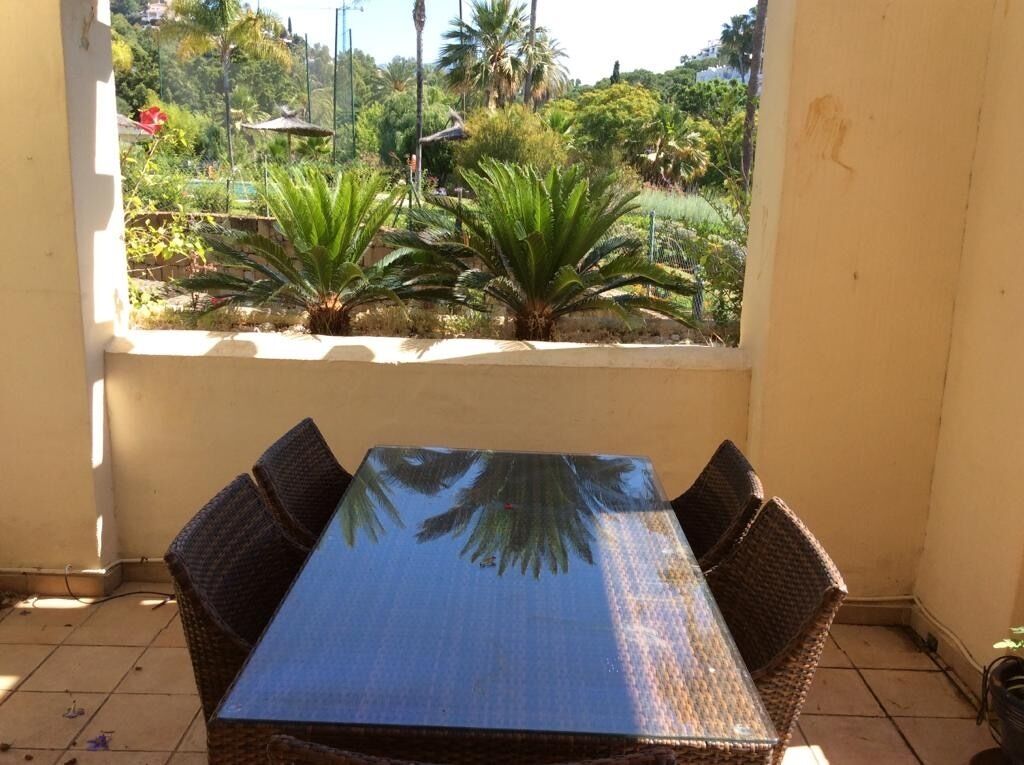 Een terras met een tafel en stoelen, uitzicht op een tuin met palmbomen, en een gebouw op de achtergrond.