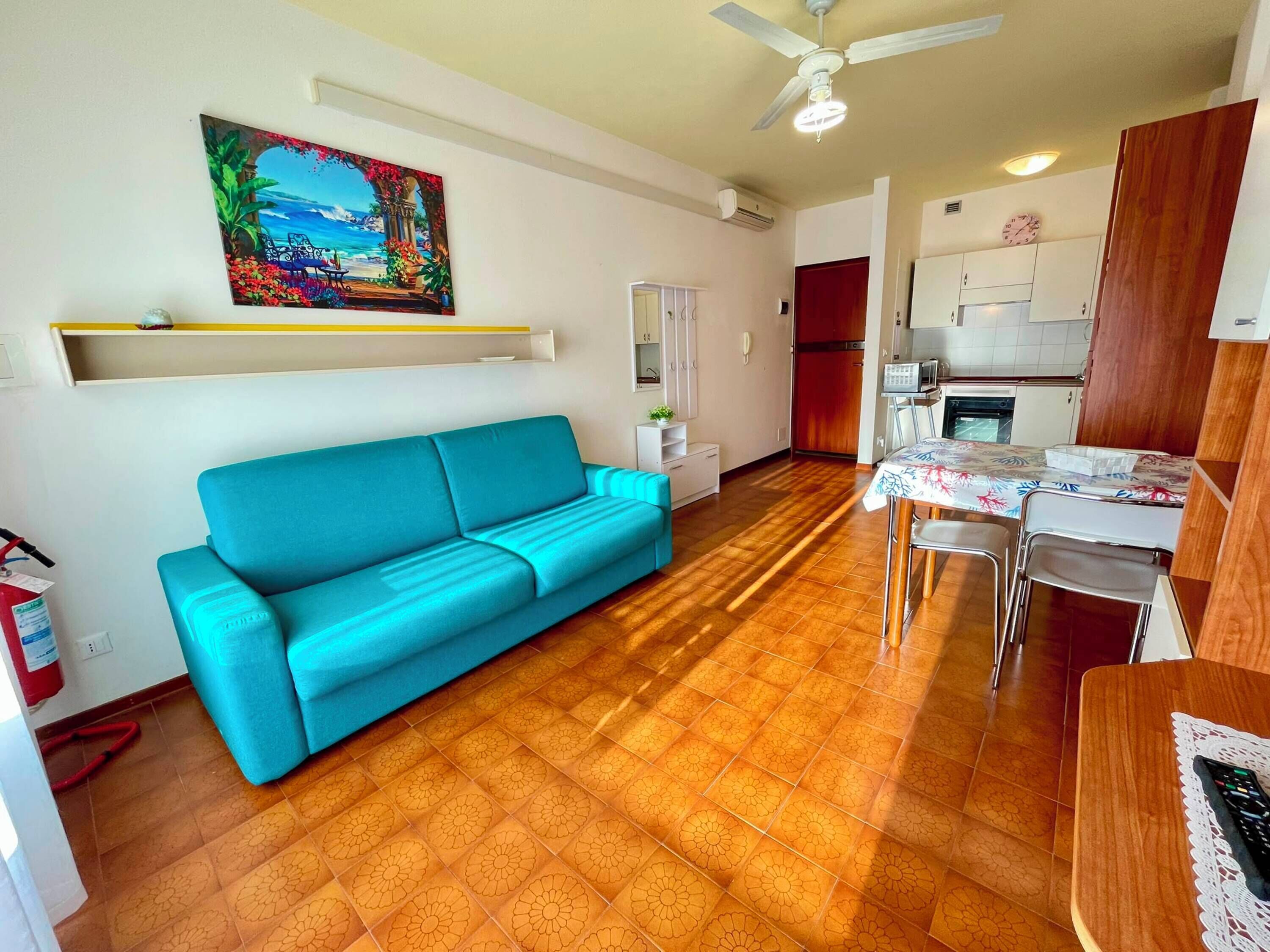 Ein Wohnzimmer mit blauem Sofa, einem Esstisch mit Stühlen, einem Deckenventilator und einer Küchenzeile mit Schränken und einem Kühlschrank.