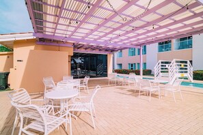 An outdoor patio area with white metal chairs and tables, a covered seating area with a purple roof, and a pool with white pool ladders.