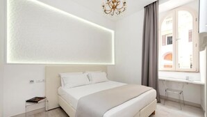 A modern bedroom with a large bed, a bedside table, a chair, a window with curtains, and a chandelier. - Primus Monti Suites  (Rome)