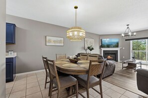 A dining area with a round table, chairs, and a chandelier. There is a fireplace with a fire, a sofa, and a coffee table.