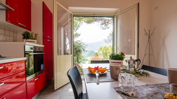 A modern kitchen with red cabinets, a dining area with a table set for two, and a view of greenery outside. - Seaside Residence with Aeolian Islands View – Pool, Gym & WiFi – Apt PANAREA (Patti)
