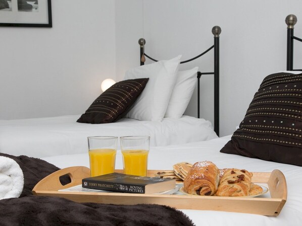 A neatly made bed with white and dark pillows, a tray with croissants and juice, and a book titled "Moving Pictures" on a wooden tray.