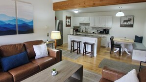 A modern living room with a brown leather sofa, a wooden coffee table, and a dining area with a round table and bar stools. - Renovated Condo, Avon Colorado, River Views and walk to Beaver Creek Shuttle (Avon)