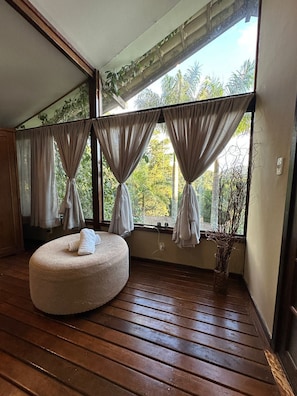 A room with a wooden floor, a round ottoman with towels, and a window with curtains. - Pousada Apoema (Petrópolis)