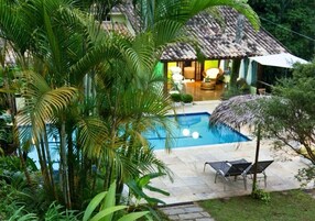 A pool area with lounge chairs, a thatched-roof structure, and lush greenery.