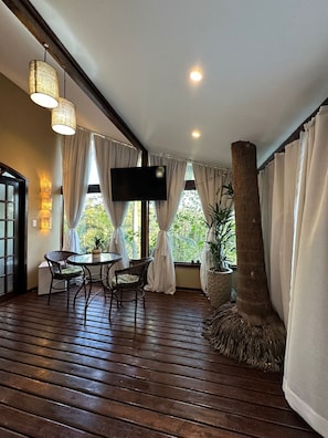 Una habitación con piso de madera, una mesa redonda con sillas, un televisor montado en la pared y ventanas amplias con cortinas.