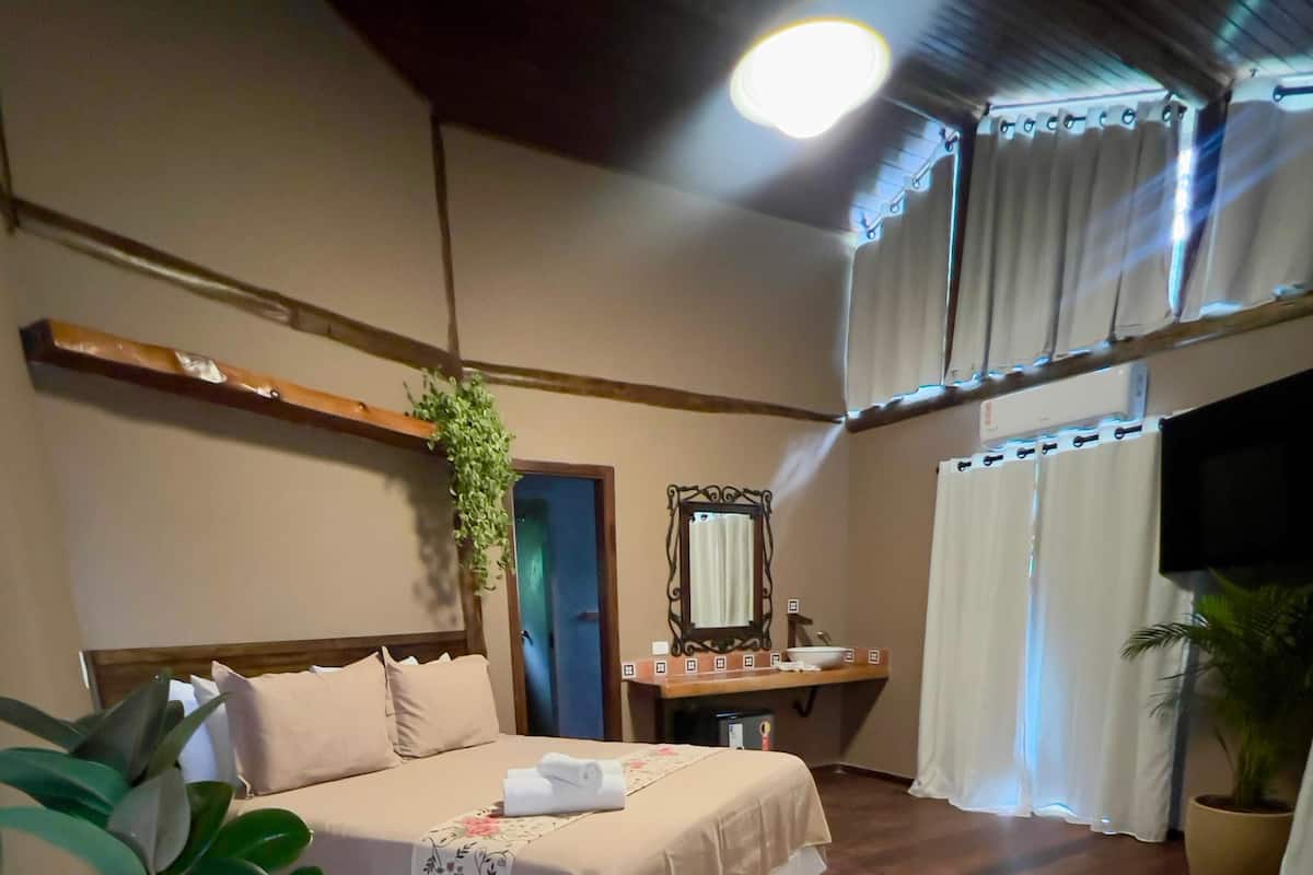 A hotel room with a bed, a bedside table, a mirror, a potted plant, and a large window with curtains.