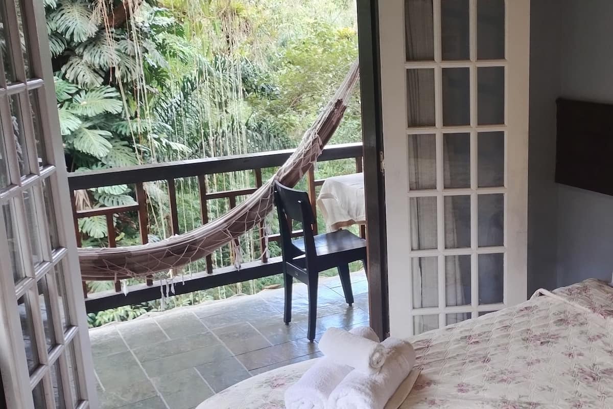 A bedroom with a bed, a hammock, and a chair on a balcony with a view of greenery.