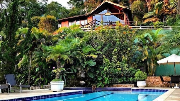 A swimming pool with a tiled edge, surrounded by lush greenery and a house in the background. - Pousada Apoema (Petrópolis)