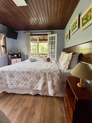 A bedroom with a wooden ceiling, a bed with a floral bedspread, a nightstand with a lamp, and a window with curtains.