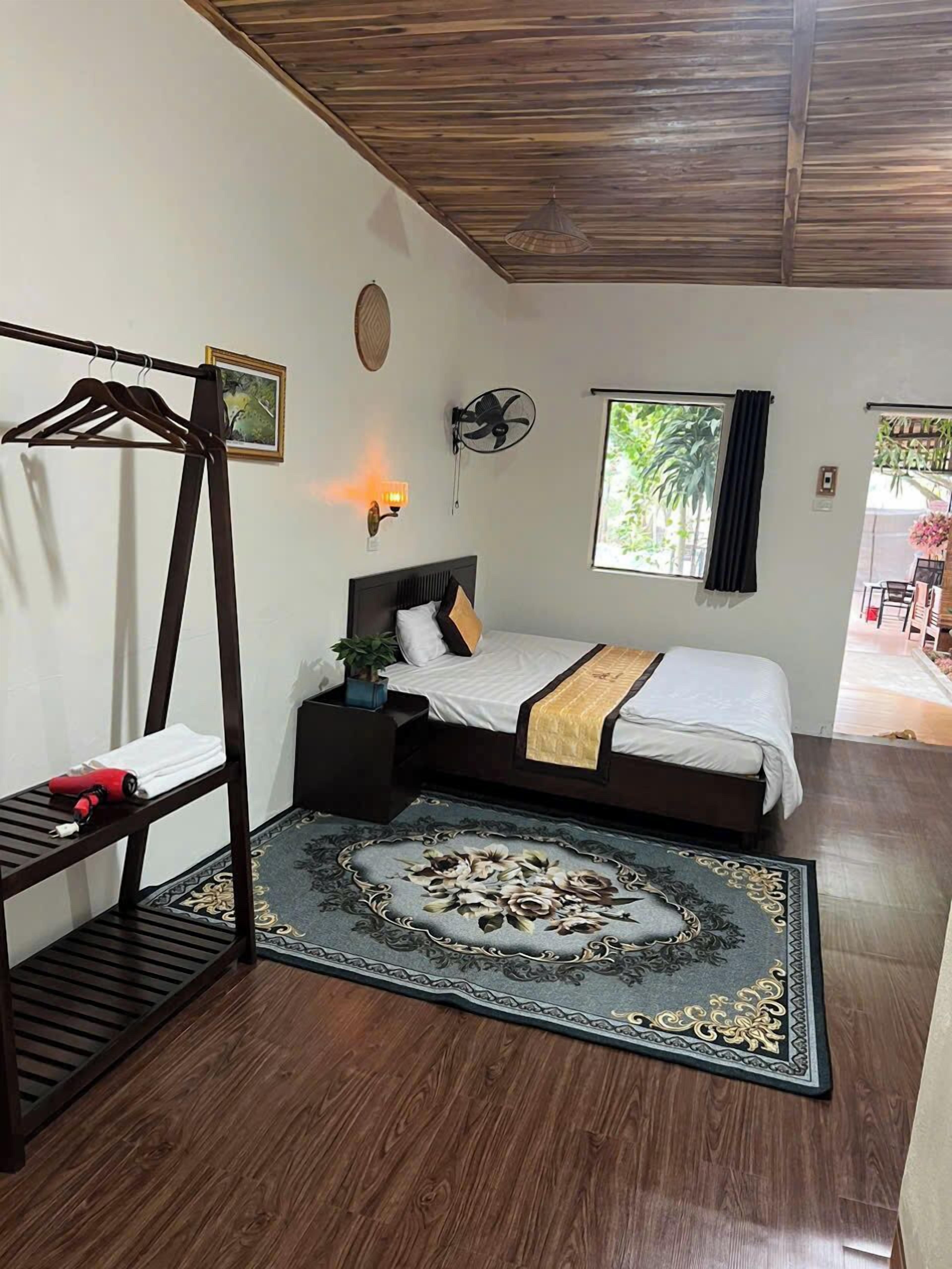 A bedroom with a bed, a wooden ceiling, a wall-mounted fan, a window with curtains, a bedside table, and a hanging rack.