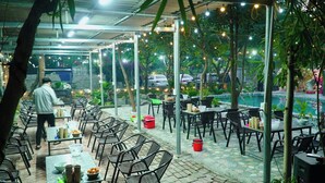 Un restaurante al aire libre con mesas y sillas, luces colgantes y vistas a una piscina y coches al fondo.