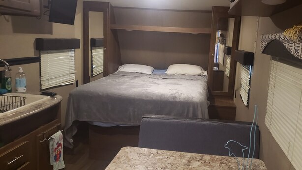 Camera da letto di un camper compatto, con un letto, un piccolo tavolo, un lavandino e una finestra con persiane.