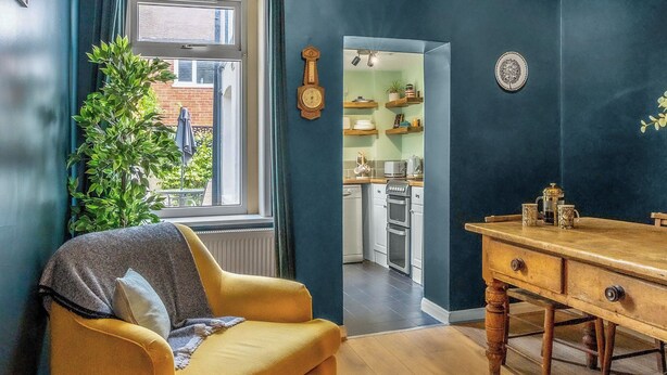 A cozy living room with a yellow armchair, a wooden dining table, and a view of a kitchen through an open door.