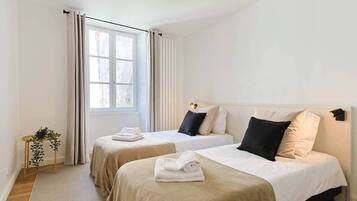 Une chambre d’hôtel avec deux lits, une fenêtre avec des rideaux, une table de chevet avec une plante et une tête de lit dotée d’un luminaire.