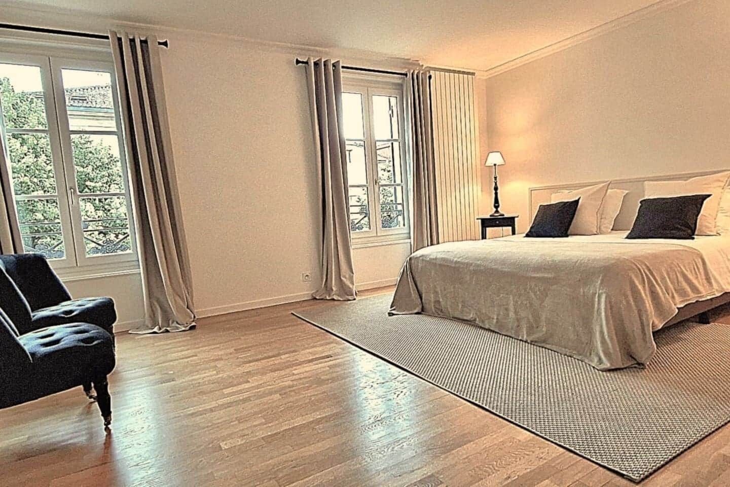 Ein Schlafzimmer mit einem Bett, einem Sessel, einer Lampe und großen Fenstern mit Vorhängen.