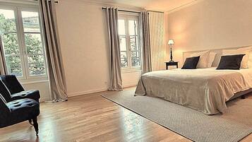 Ein Schlafzimmer mit einem Bett, einem Sessel, einer Lampe und großen Fenstern mit Vorhängen.