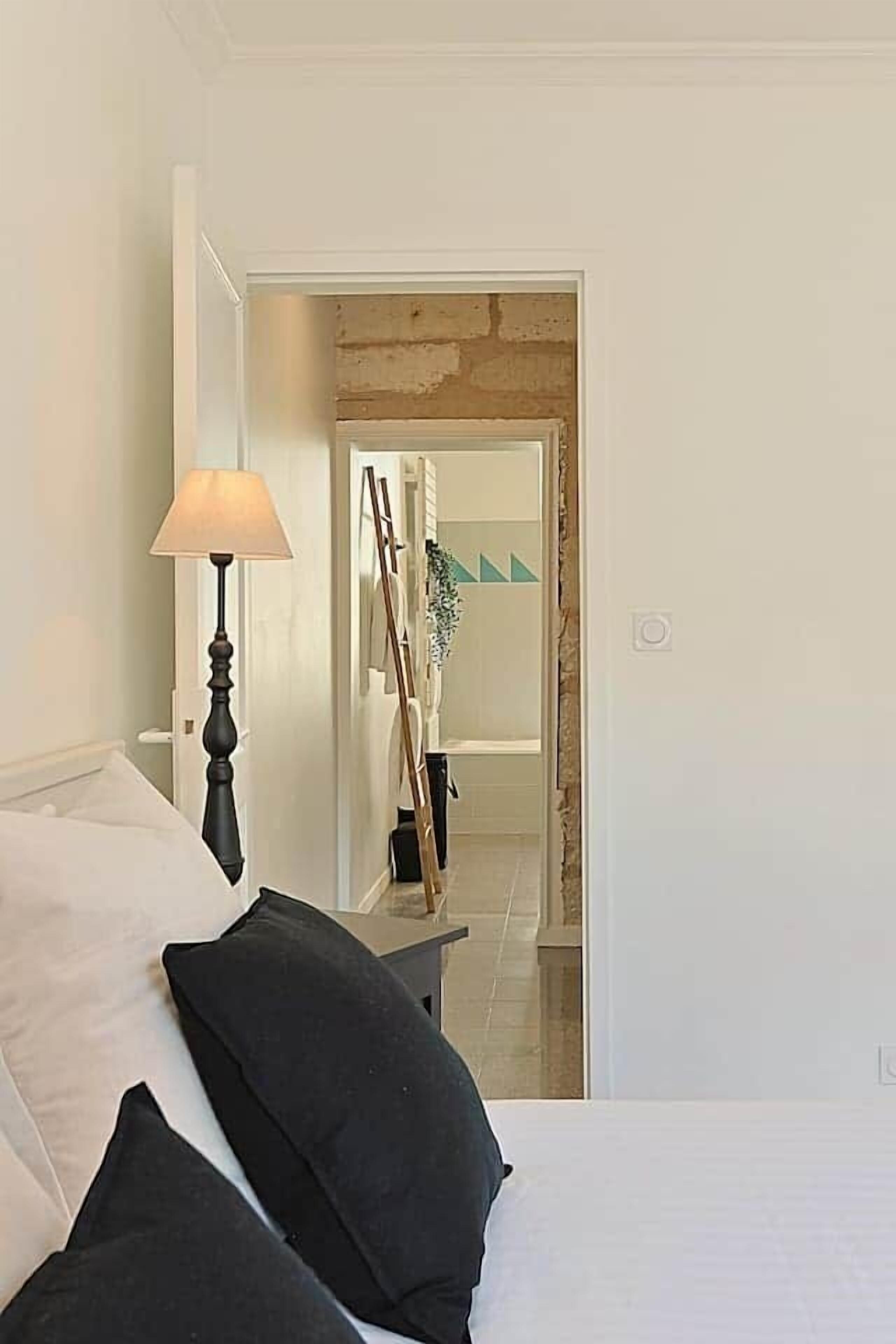 Et soveværelse med seng, lampe og udsigt til et badeværelse med spejl og håndklædestang.