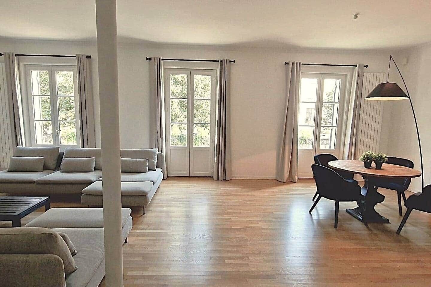Een ruime woonkamer met een zithoek, een eettafel met stoelen en grote ramen met gordijnen.
