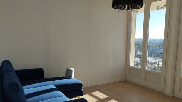 Ett rum med en blå soffa, en liten vit högtalare och ett stort fönster med utsikt över en stad.