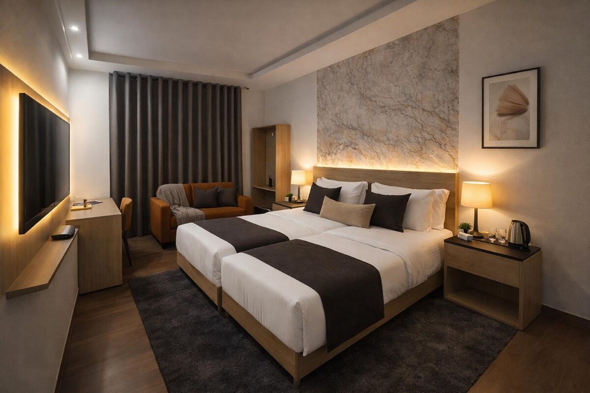 A modern hotel room with a large bed, bedside tables, a desk, a chair, a TV, and a marble wall.