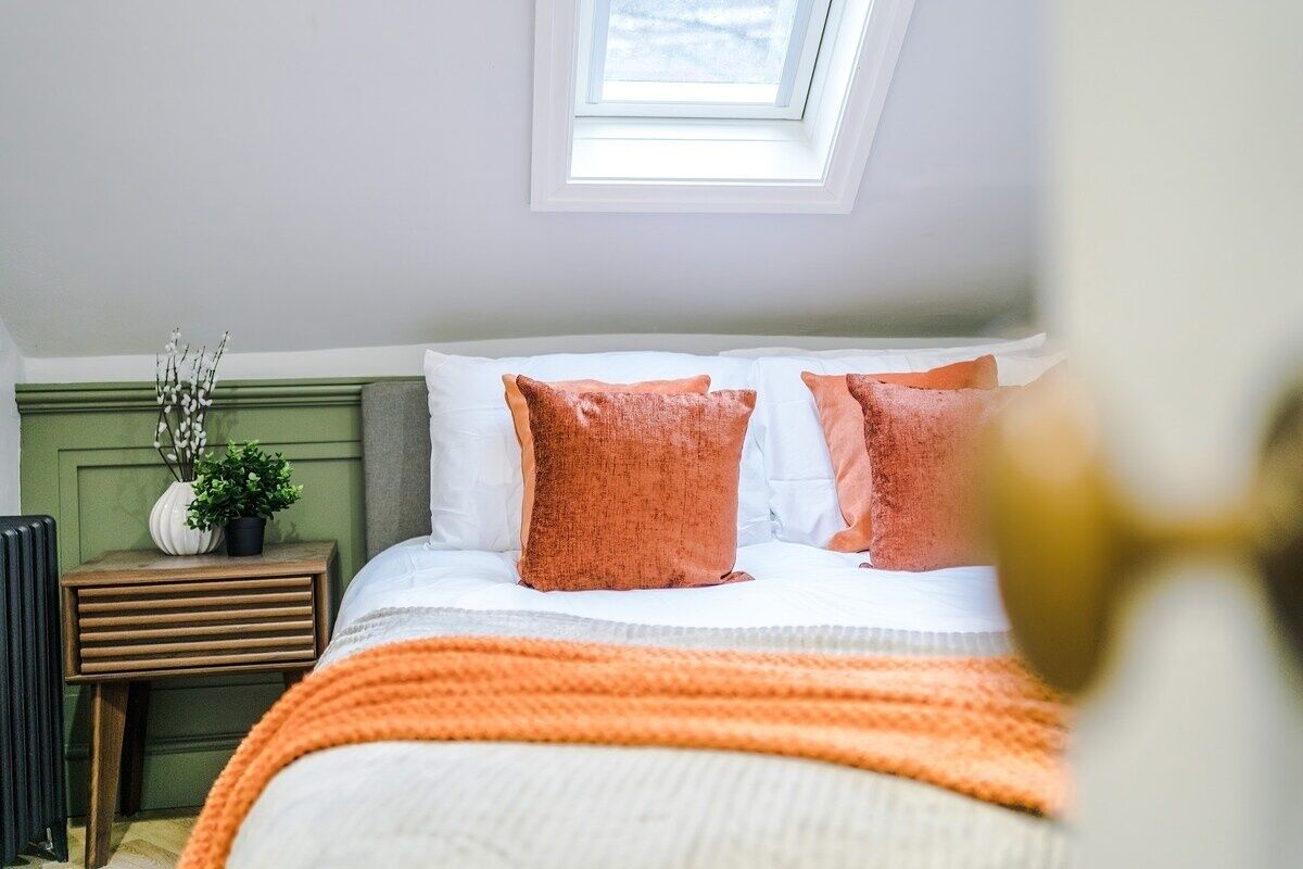 A bedroom with a skylight, a bed with white and orange bedding, a bedside table with a plant, and a radiator.