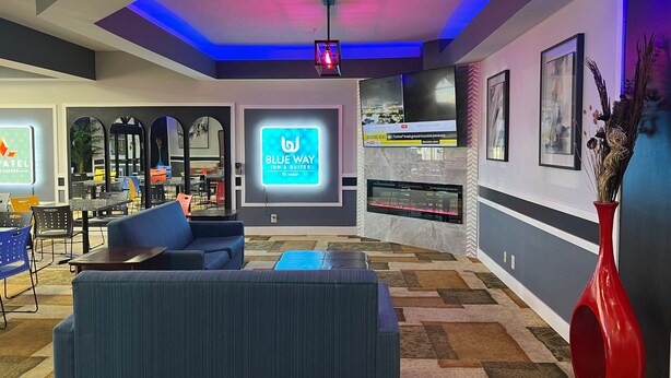 Un hall d'hôtel moderne avec un canapé bleu, une cheminée et un panneau lumineux néon indiquant « Blue Way Suites ».