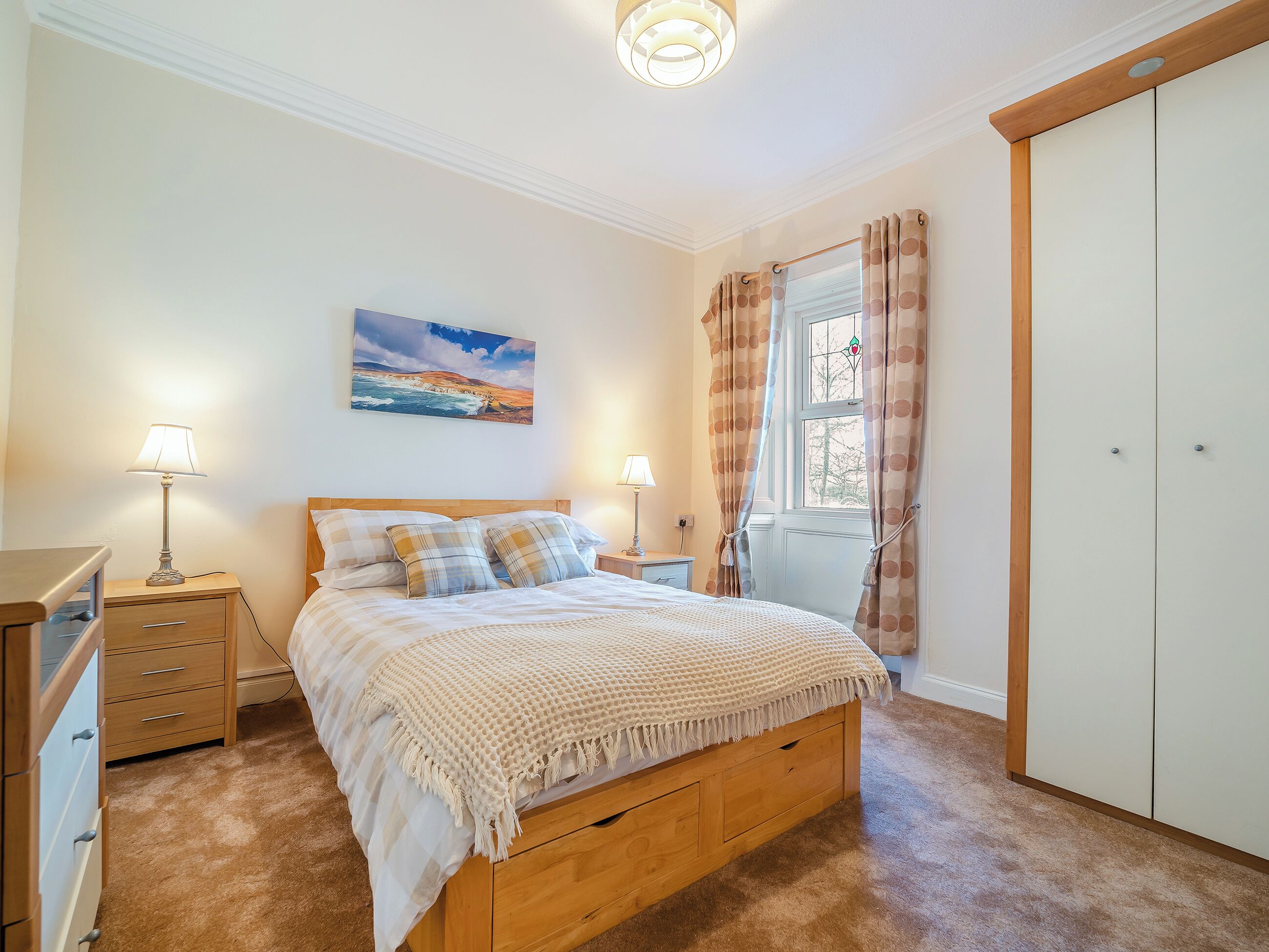 A bedroom with a wooden bed, two bedside tables with lamps, a large window with curtains, a wardrobe, and a painting on the wall.