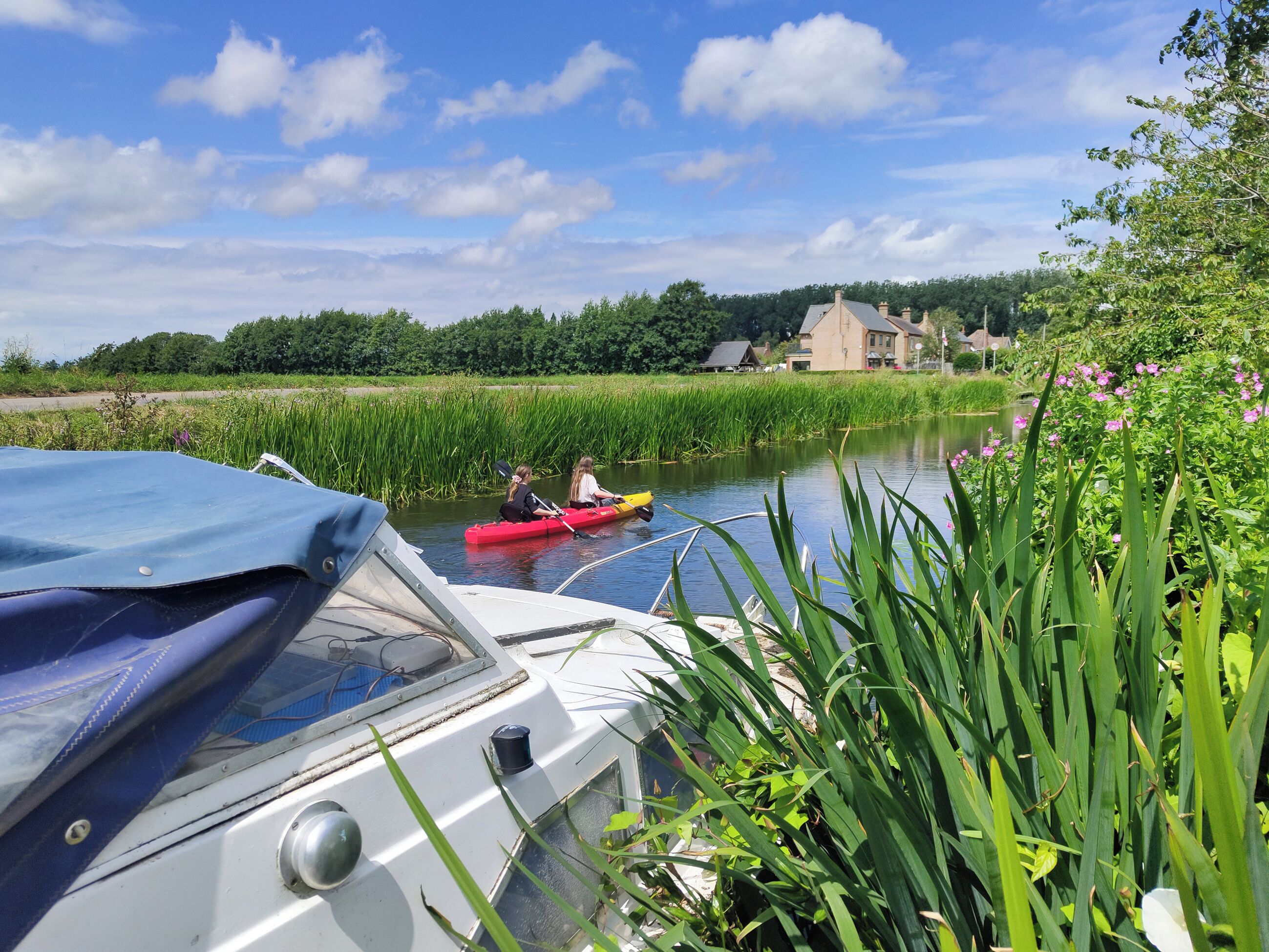 A boat docked near a river with two people in a red kayak.