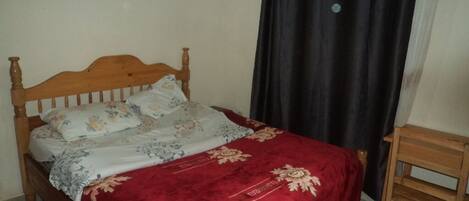 Una cama con cabecera de madera, colcha estampada con motivos florales y una manta roja. Junto a la cama hay una pequeña mesita de madera.