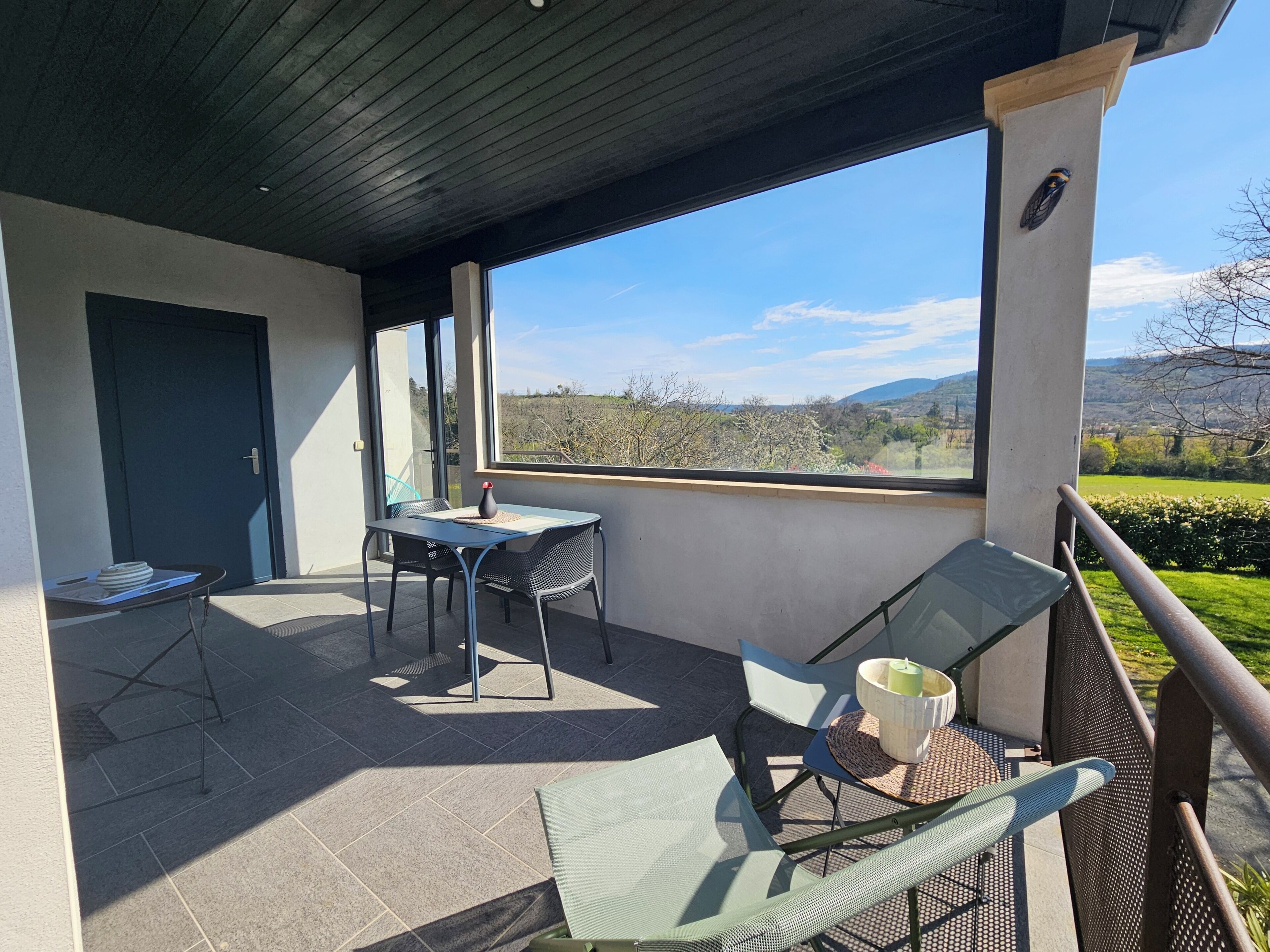 Ein überdachter Außenbereich mit Tisch und Stühlen, einer Tür und Blick auf die Landschaft.