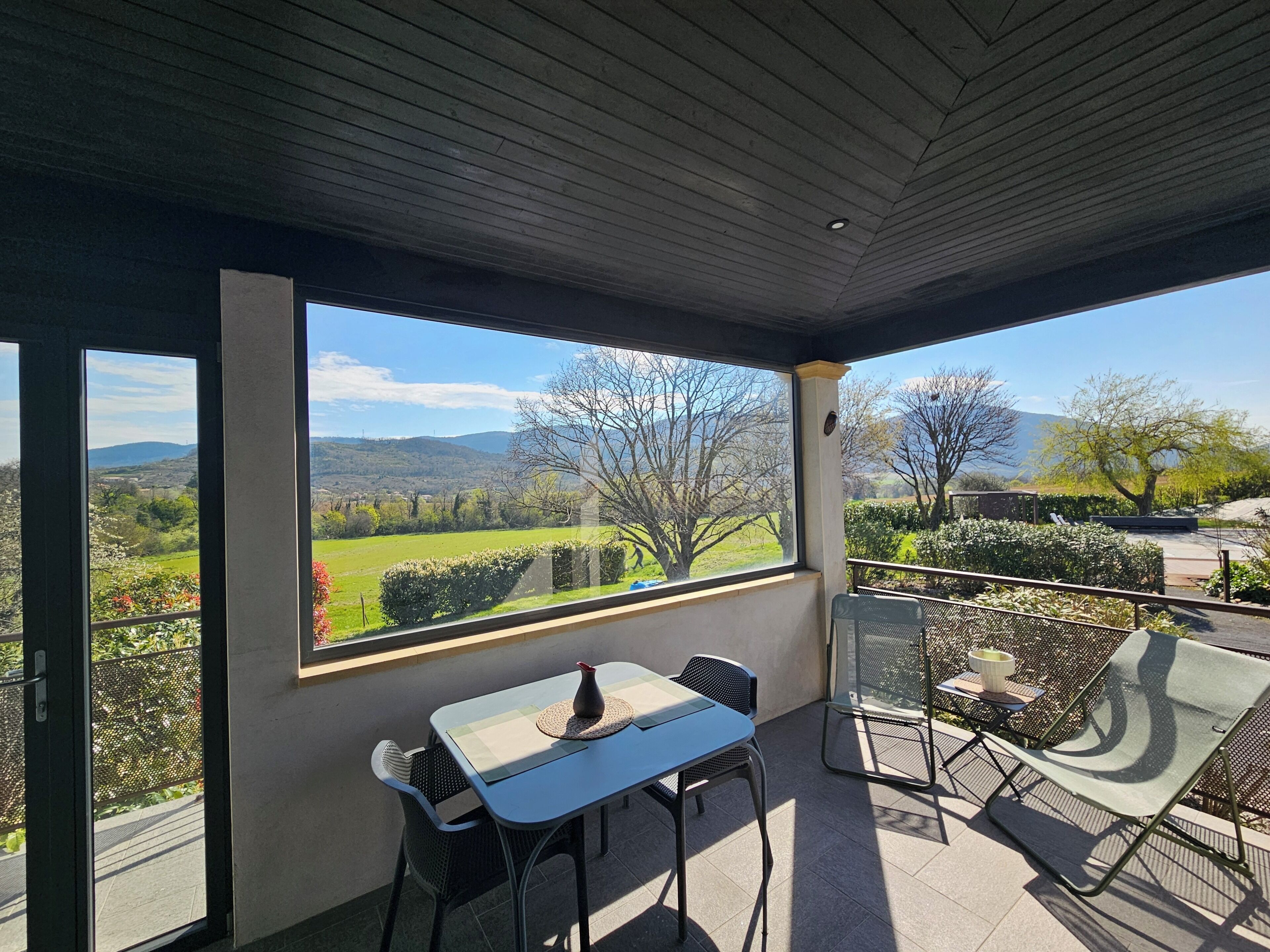 A covered patio with a table and chairs, large windows offering a view of a green landscape.