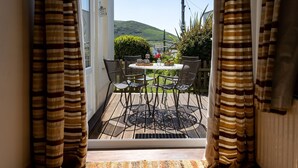 Une vue depuis une fenêtre ornée de rideaux à rayures, donnant sur un patio extérieur avec une table et des chaises, un petit jardin et une colline en arrière-plan.