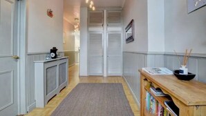 Un couloir doté d’un plancher en bois, d’un tapis gris, d’une console en bois sur laquelle sont posés des livres, d’une petite table noire avec une horloge, et de portes blanches avec des volets.
