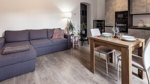 Una sala de estar moderna con un sofá gris, una mesa de comedor de madera y una cocina equipada con microondas y horno.