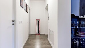 Un pasillo con suelo de madera, paredes blancas y cuadros en la pared izquierda. Un espejo con marco rojo se encuentra en el centro, y un aparador de cristal con copas de vino a la derecha.
