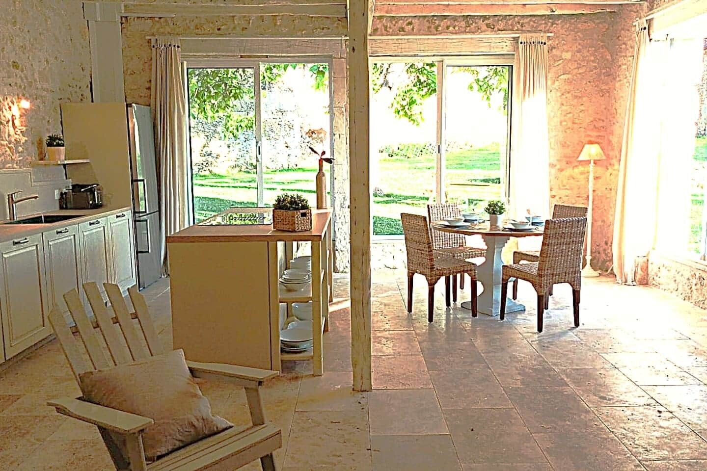 Een keuken met witte kastjes, een eethoek met rieten stoelen, en een terras met een tafel en stoelen.
