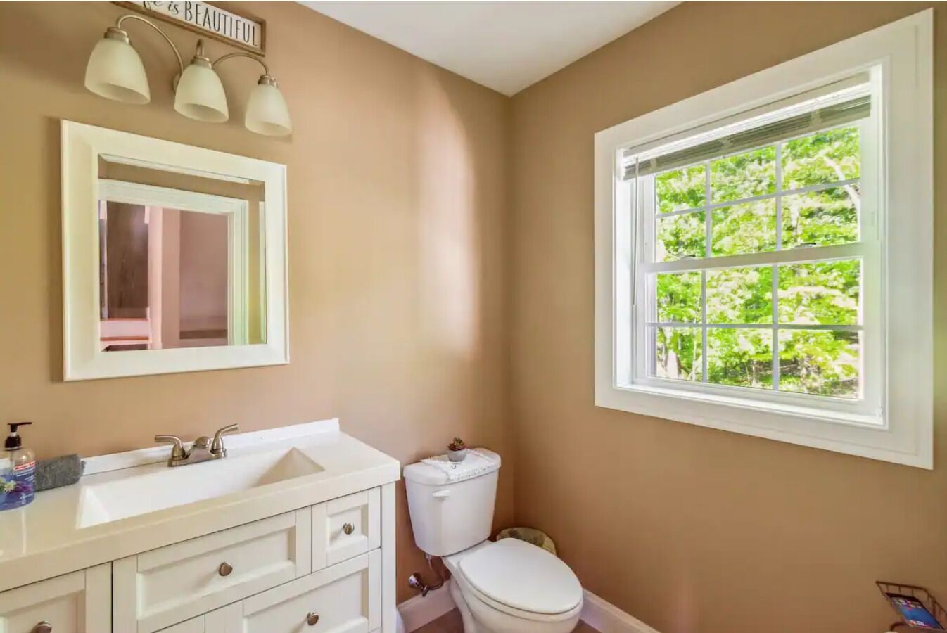 Een badkamer met een beige muur, een wit wastafelmeubel met een spiegel erboven, een toilet en een raam met uitzicht op groen.