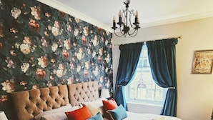 A bedroom with a tufted headboard, floral wallpaper, a chandelier, and a window with curtains. - Saltaire Retreat 3 Bed House (Shipley)