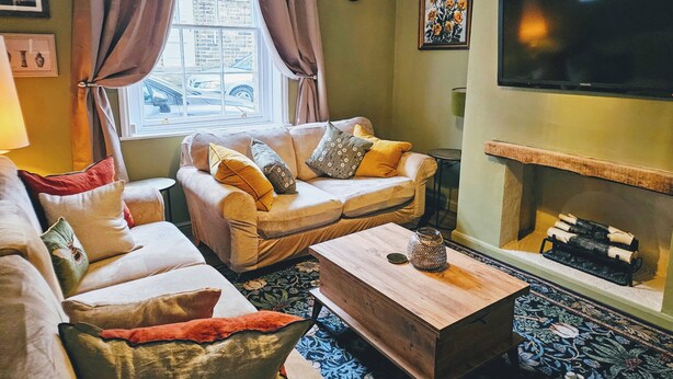 A cozy living room with a sofa, a coffee table, and a TV. There's a window with curtains, a clock, and a painting on the wall. - Saltaire Retreat 3 Bed House (Shipley)