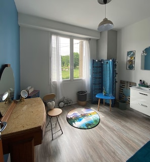 A room with a large window, a round table with a scenic view, a blue chair, a small bicycle, and a white cabinet.