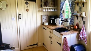 A cozy kitchen with a wooden door labeled "WASHROOM," a coffee maker, and a kettle on the counter. - Lilly's Pad (Bude)