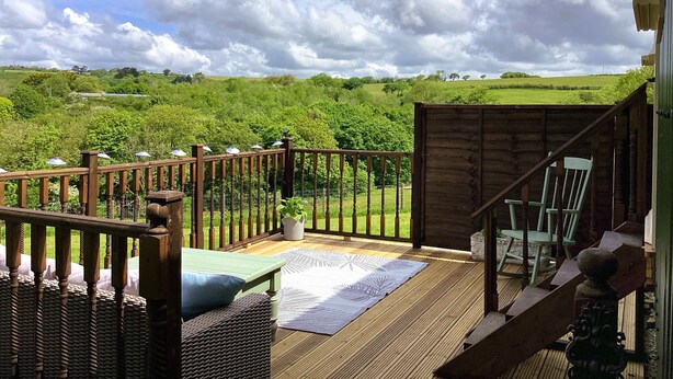 A wooden deck with a view of a green landscape, a wicker sofa, and a chair. - Lilly's Pad (Bude)