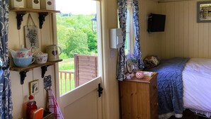 A cozy room with a wooden door leading to a balcony, a bed with a blue blanket, a shelf with kitchenware, and a wall-mounted TV. - Lilly's Pad (Bude)