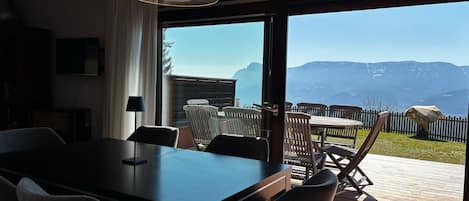 A dining area with a table and chairs, a television, and a view of mountains through a large glass door.